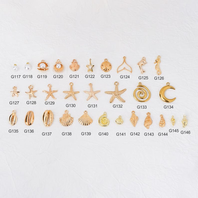May include: A collection of gold-colored charms in various shapes, including seashells, starfish, pearls, and a crescent moon. The charms are arranged on a white surface and labeled with individual codes.