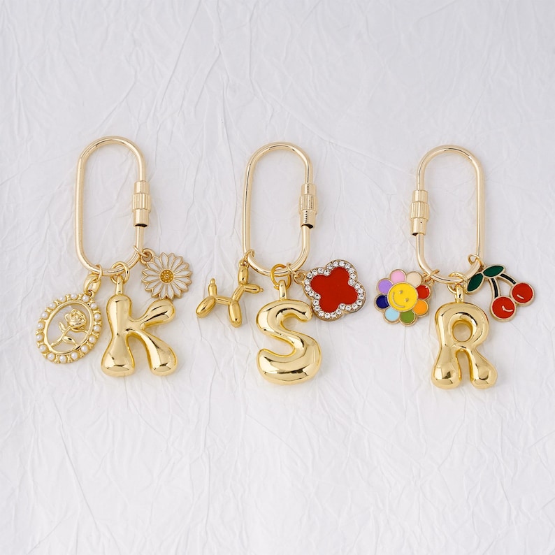 May include: Gold-colored keychains with various charms. One keychain has the letter "K" with a daisy and an oval charm. Another has the letter "S" with a balloon dog and a red flower. The last has the letter "R" with a flower and cherries.