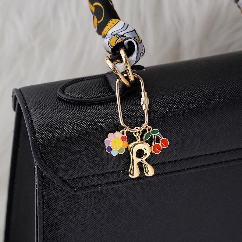 May include: A black handbag with a gold-colored charm. The charm features a colorful flower, the letter "R", and cherries. The handbag's handle is wrapped with a patterned scarf. The charm is attached to the bag with a gold carabiner.