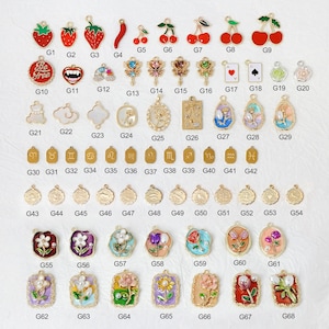 May include: A collection of gold-toned charms and pendants with various designs, including fruits, playing card suits, flowers, and zodiac symbols. Some charms feature enamel detailing in red, green, and blue. The charms are suitable for jewelry making.