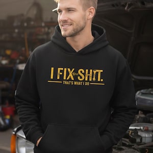 May include: Black hooded sweatshirt with the text "I FIX SHIT. THAT'S WHAT I DO" in yellow. The sweatshirt has a front pocket and a drawstring hood. The person in the image is wearing blue jeans.