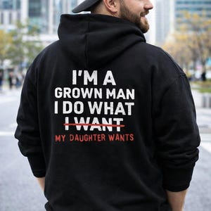 May include: Black hooded sweatshirt with white and red text: "I'M A GROWN MAN I DO WHAT I WANT MY DAUGHTER WANTS." The sweatshirt is worn by a person wearing a black baseball cap.