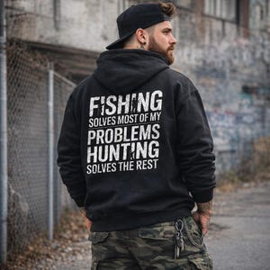 May include: Black hooded sweatshirt with white text: "FISHING SOLVES MOST OF MY PROBLEMS HUNTING SOLVES THE REST." The sweatshirt is worn with camouflage cargo pants and a black baseball cap. The image is taken outdoors.