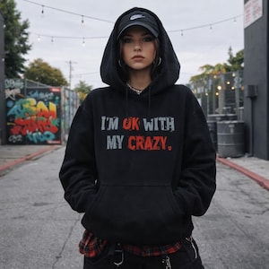 May include: Black hoodie with the text "I'M OK WITH MY CRAZY." in red and gray. The hoodie has a drawstring hood and a front pocket. A black baseball cap is worn on the head.