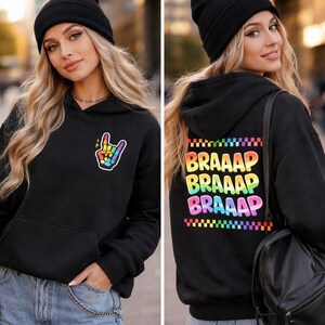 May include: Black hoodie with a rainbow hand gesture graphic on the front and the word "BRAAAP" in rainbow letters on the back. The hoodie has a front pocket and a hood. The model is wearing a black beanie and blue jeans.