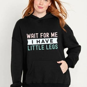 May include: Black hooded sweatshirt with the text "WAIT FOR ME I HAVE LITTLE LEGS" in white, pink, and teal. The sweatshirt has a front pocket and black cuffs.