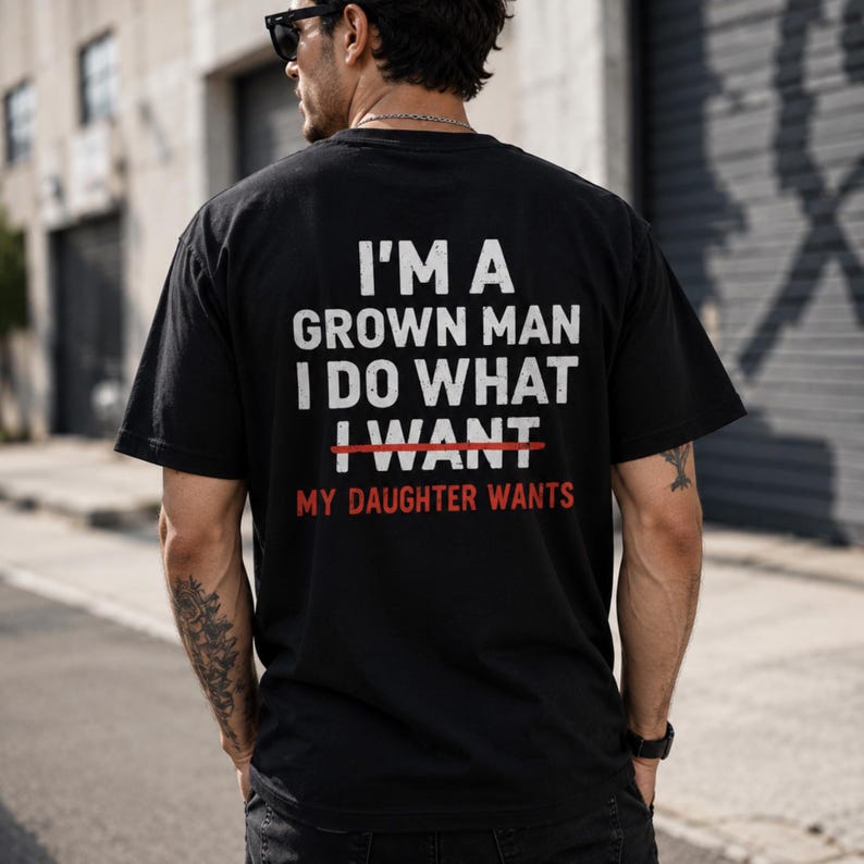 Puede incluir: Camiseta negra con texto blanco en la espalda. El texto dice "I'M A GROWN MAN I DO WHAT I WANT, MY DAUGHTER WANTS". La palabra "WANT" est&aacute; tachada en rojo. Prenda de vestir informal.