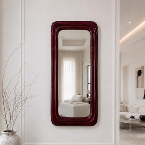 May include: A rectangular wall mirror featuring a deep, glossy burgundy frame with rounded corners. The mirror has a smooth, reflective surface that displays a clear view of the interior space, including a white sofa and sheer curtains.