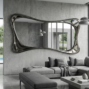 Può includere: Un grande specchio dalla forma irregolare con una cornice metallica è appeso a una parete di cemento. Lo specchio riflette un moderno soggiorno con un divano grigio, un tavolino e grandi finestre. Il design è contemporaneo.