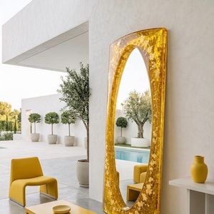May include: A large, gold-colored mirror with an oval shape and gold leaf accents stands against a white wall. The mirror reflects an outdoor scene with trees and a pool. Yellow furniture and a yellow vase are also visible.