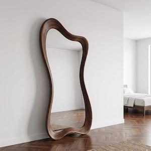 May include: A large, free-form wooden mirror leans against a white wall. The mirror's frame is a rich, dark brown with a wavy, organic shape. The mirror reflects the room, including a bed and a window. The floor is made of wood.