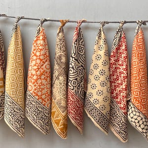 May include: A collection of ten patterned fabric bandanas hanging from a thin, dark-colored wire. The bandanas feature various designs in shades of orange, red, brown, and cream, with geometric and floral patterns. The bandanas are knotted at the top.