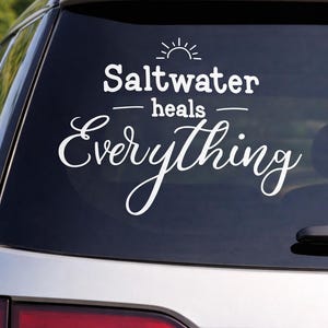 May include: White vinyl decal applied to a car window. The decal features the phrase "Saltwater heals Everything" in a stylized font, with a sun graphic above the word "Saltwater".