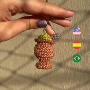 Pode incluir: Chaveiro de crochê feito à mão com um vaso marrom com uma parte superior verde. O chaveiro é preso a um anel de metal prateado. O fundo é de cor bege neutra. O chaveiro é segurado por uma mão.