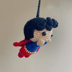 Pode incluir: Uma figura do Super-Homem de crochê feita à mão em voo. A figura tem um rosto bronzeado, cabelo preto e usa uma fantasia azul e vermelha com um emblema amarelo. Uma capa vermelha está presa nas costas. A figura é suspensa por um fio azul escuro.