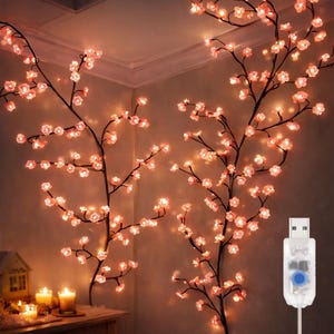 May include: Cherry blossom branch lights with pink flowers and warm white LED lights. The branches are mounted on a wall, creating a soft, ambient glow. A USB connector is visible. Candles and a small house are on a table.
