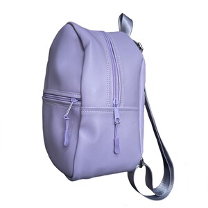 Könnte beinhalten: Ein lila-farbener Rucksack mit einem Reißverschluss-Hauptfach und einer kleineren Reißverschluss-Vordertasche. Die Tasche hat einen oberen Griff und verstellbare Schultergurte. Die Reißverschlüsse und Zipper sind farblich passend zur Tasche.