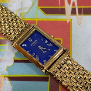 May include: A gold-toned Seiko Quartz watch with a rectangular blue face and gold Roman numerals. The watch has a woven gold band. The watch face has the Seiko Quartz logo. The watch is a classic design.
