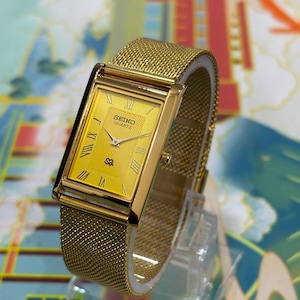 May include: A gold-toned Seiko Quartz watch with a rectangular face and mesh band. The watch face is yellow with Roman numerals and the Seiko Quartz logo. The watch is displayed on a clear stand.