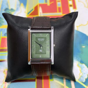 Puede incluir: Un reloj Seiko Quartz rectangular con esfera verde y caja plateada, sobre un cojín negro. El reloj presenta números romanos y una correa de cuero marrón. La esfera del reloj tiene el logotipo de Seiko Quartz.