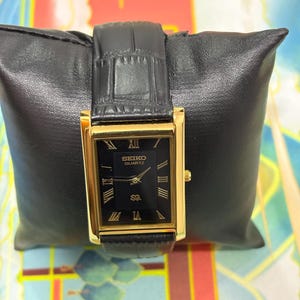 May include: A rectangular Seiko Quartz watch with a black face and gold-tone case and hands. The watch features Roman numeral hour markers and a black leather band. The watch is displayed on a black leather pillow.