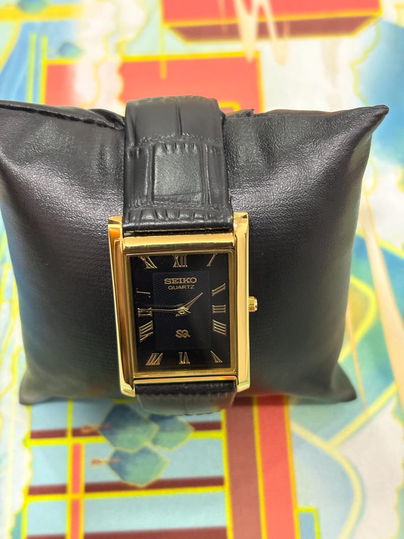 May include: A rectangular Seiko Quartz watch with a black face and gold-tone case and hands. The watch features Roman numeral hour markers and a black leather band. The watch is displayed on a black leather pillow.