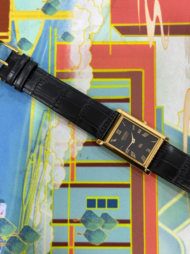 May include: A rectangular gold-toned wristwatch with a black face and Roman numeral hour markers. The watch has a black leather band with a buckle closure. The watch face reads "SEIKO QUARTZ".