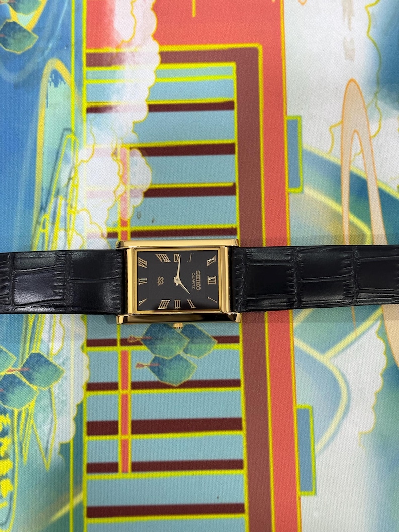 May include: A rectangular gold-toned watch with a black face and black leather band. The watch face features gold Roman numerals and the text "SEIKO QUARTZ". The watch band is textured with a crocodile pattern.