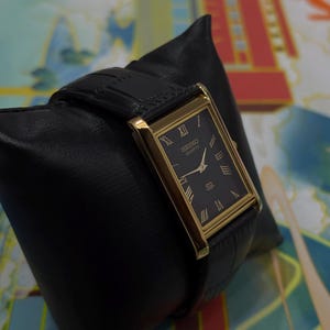 May include: A rectangular Seiko Quartz watch with a black face and gold-colored frame. The watch has Roman numeral hour markers and a black leather band. The watch is displayed on a black leather pillow.
