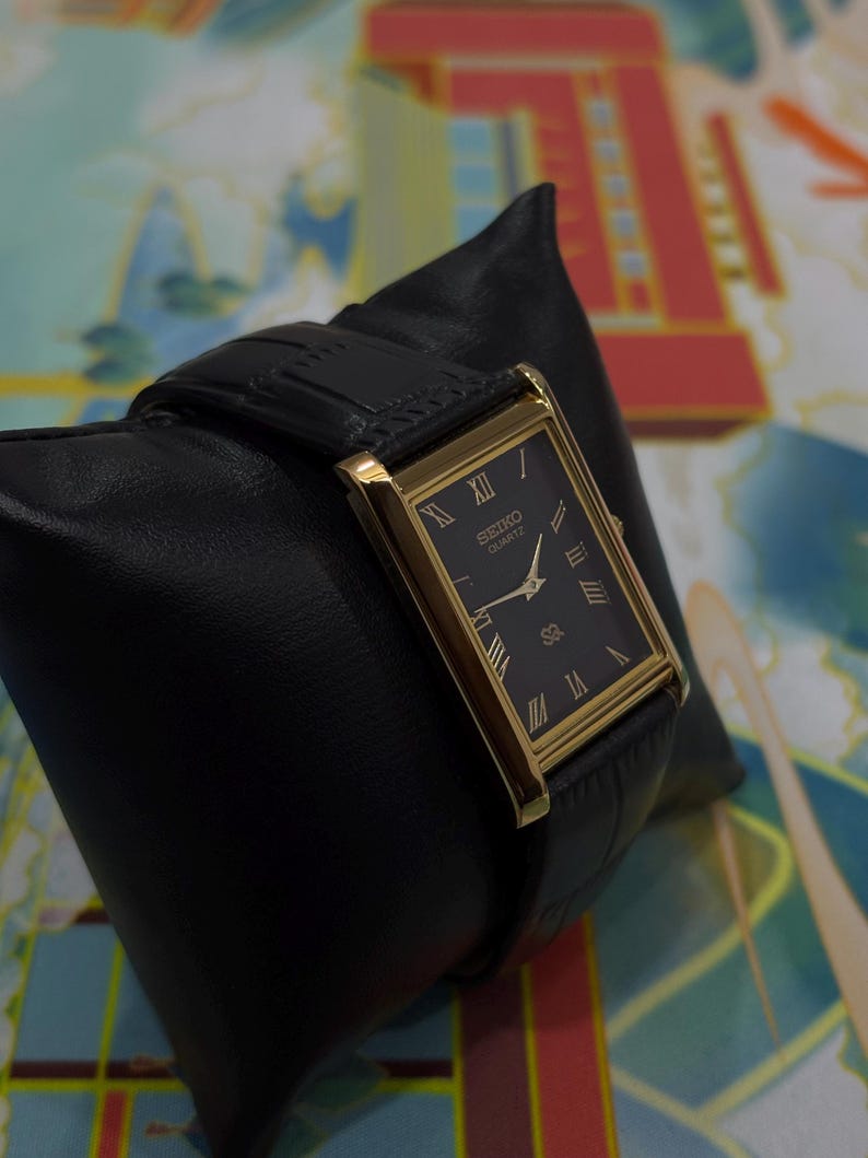 May include: A rectangular Seiko Quartz watch with a black face and gold-colored frame. The watch has Roman numeral hour markers and a black leather band. The watch is displayed on a black leather pillow.