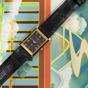 May include: A rectangular gold-toned Seiko Quartz watch with a black face and Roman numeral hour markers. The watch has a black leather band with a crocodile pattern. The watch is displayed on a colorful patterned surface.