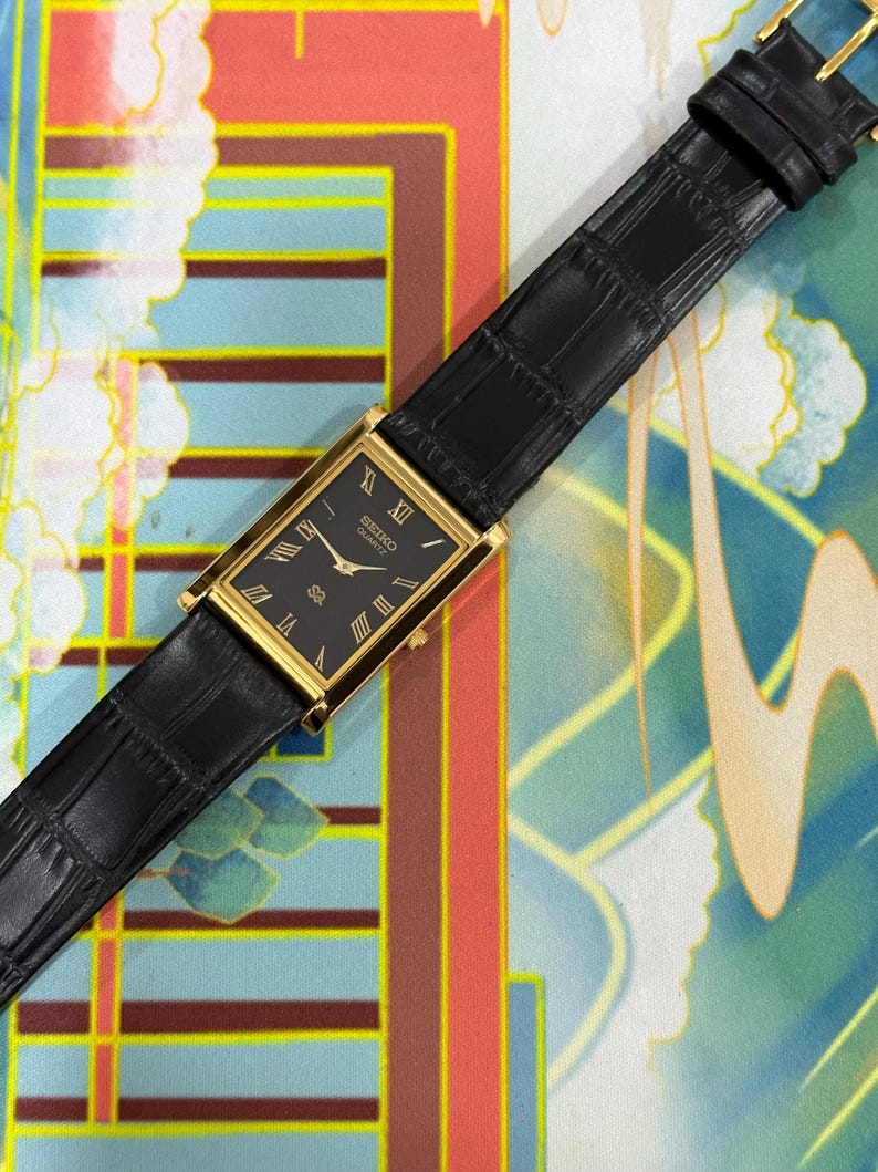 May include: A rectangular gold-toned Seiko Quartz watch with a black face and Roman numeral hour markers. The watch has a black leather band with a crocodile pattern. The watch is displayed on a colorful patterned surface.