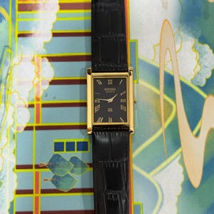 May include: A rectangular Seiko Quartz watch with a black face and gold-colored case. The watch has a black leather band. Roman numerals mark the hours. The watch is a classic design.
