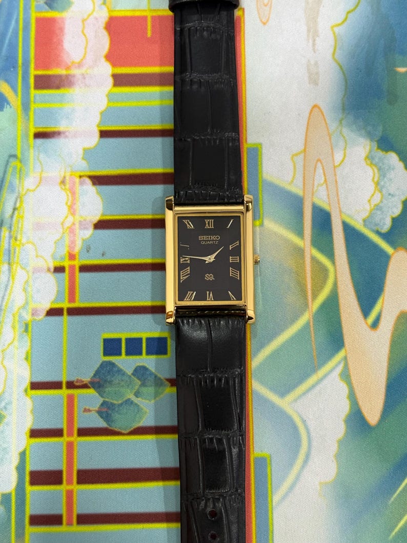 May include: A rectangular Seiko Quartz watch with a black face and gold-colored case. The watch has a black leather band. Roman numerals mark the hours. The watch is a classic design.