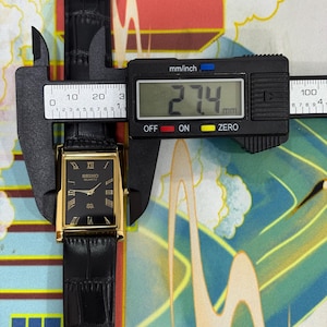 May include: A rectangular Seiko Quartz watch with a black dial, gold-colored case, and black leather band. The watch is being measured with a digital caliper, showing a width of 27.4 mm. The watch face features Roman numerals and the Seiko Quartz logo.