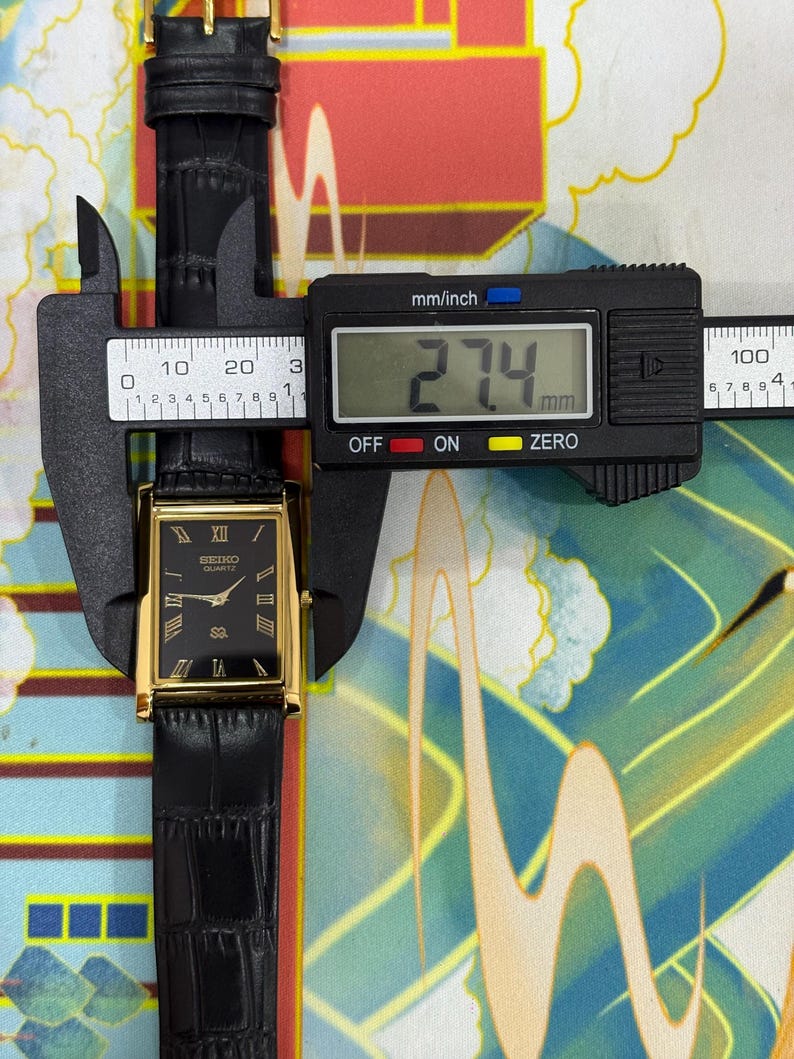 May include: A rectangular Seiko Quartz watch with a black dial, gold-colored case, and black leather band. The watch is being measured with a digital caliper, showing a width of 27.4 mm. The watch face features Roman numerals and the Seiko Quartz logo.