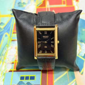 May include: A rectangular gold-toned Seiko Quartz watch with a black face and black leather band. The watch face displays Roman numerals and the Seiko Quartz logo. The watch is displayed on a black leather pillow.
