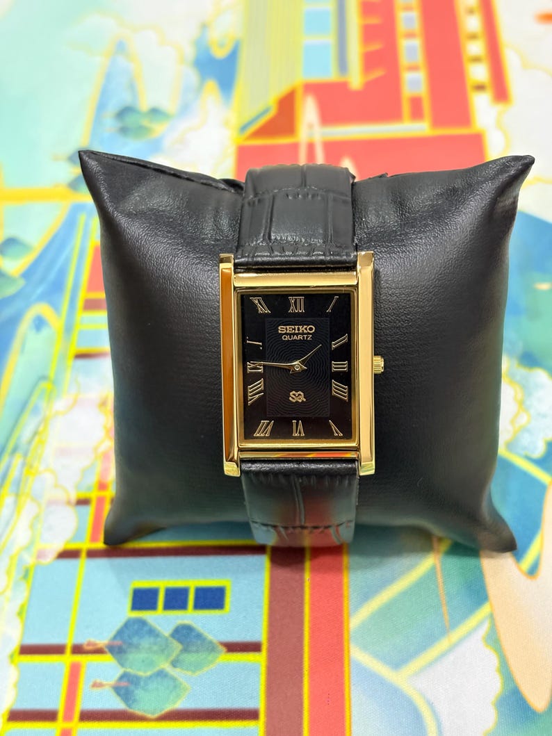 May include: A rectangular gold-toned Seiko Quartz watch with a black face and black leather band. The watch face displays Roman numerals and the Seiko Quartz logo. The watch is displayed on a black leather pillow.