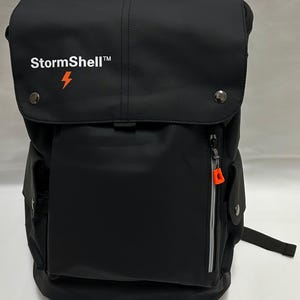 May include: Black backpack with a flap closure and multiple pockets. The backpack features the text "StormShell" and a lightning bolt logo. It has a zippered front pocket with an orange zipper pull and silver accents.