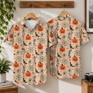 May include: A short-sleeved button-down shirt in a light beige color featuring an all-over Halloween print. The pattern includes orange jack-o'-lanterns, black spiderwebs, black spiders, black bats, and dancing skeletons on a light background.