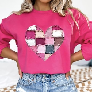 Può includere: Una felpa rosa acceso con un design a cuore patchwork. Il cuore è composto da quadrati di tessuto in tonalità rosa, bianco e marrone, cuciti insieme con filo visibile. La felpa ha maniche lunghe e scollo tondo.