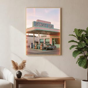 May include: Framed print of a vintage gas station in pink and teal. The sign reads "Sunset Gas" and "Coffee". A classic car is parked beneath the canopy. The image has a retro aesthetic, evoking a sense of nostalgia.