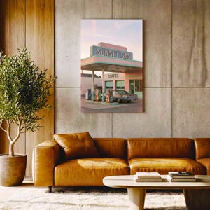 May include: A framed print depicting a vintage gas station, "Sunset Gas", in pastel pink and teal. The image shows a classic car at the pumps, with the word "Coffee" visible. The print is displayed above a brown leather sofa and a round wooden coffee table.
