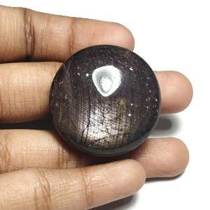 Könnte beinhalten: Ein polierter, kugelförmiger Edelstein mit einer dunklen, schillernden Oberfläche. Der Stein hat eine Mischung aus dunklen Lila- und Silbertönen mit einem hellen, reflektierenden Bereich in der Nähe der Oberseite. Die Kugel wird in einer Hand gehalten.