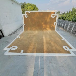 May include: A large, rectangular rug with a tan center and a white border. The border features decorative, curved white accents. The rug is displayed outdoors, with a concrete surface and a clear sky in the background.