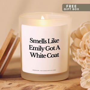 May include: A frosted glass candle in a jar with a lit wick and a bamboo lid. The white label features black text that reads Smells Like Emily Got A White Coat and Gilded Glow 8oz Natural Soy Wax Candle. The candle is lit and glowing.