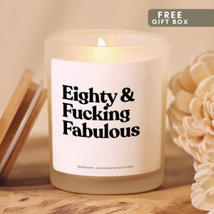 May include: A frosted glass candle with a bamboo lid, featuring a white label with the black text Eighty & Fucking Fabulous and Gilded Glow 8 oz natural soy wax candle. The candle is lit, emitting a warm glow, and is positioned next to a wooden flower.