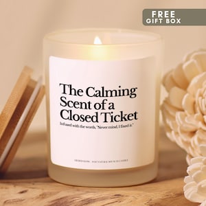 May include: A frosted glass candle with a wooden lid, featuring a white label with black text that reads: The Calming Scent of a Closed Ticket. Infused with the words, Never mind, I fixed it. GILDED GLOW | 8OZ NATURAL SOY WAX CANDLE.
