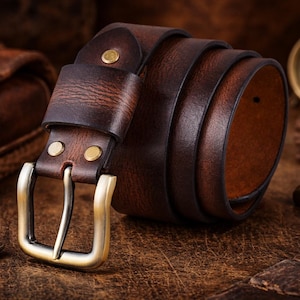 Può includere: Una cintura in pelle marrone arrotolata con fibbia e rivetti in ottone. La cintura ha un aspetto usurato e strutturato, che suggerisce una lavorazione di qualità. La fibbia è un design classico e l'estetica generale è rustica e durevole.