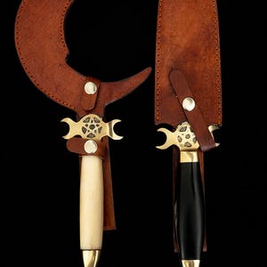 Puede incluir: Dos cuchillos artesanales con fundas de cuero. Uno tiene una hoja curva y un mango de hueso, el otro una hoja recta y un mango negro. Ambos presentan detalles en latón y forma de media luna.